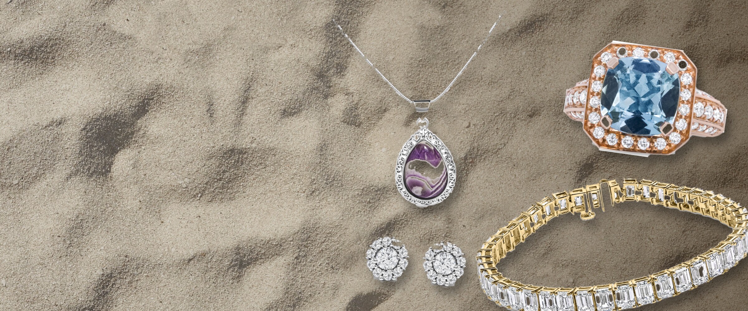 Fine jewelry arranged on beach sand: an aquamarine rose gold ring, a diamond tennis bracelet, an amethyst pendant necklace and diamond stud earrings